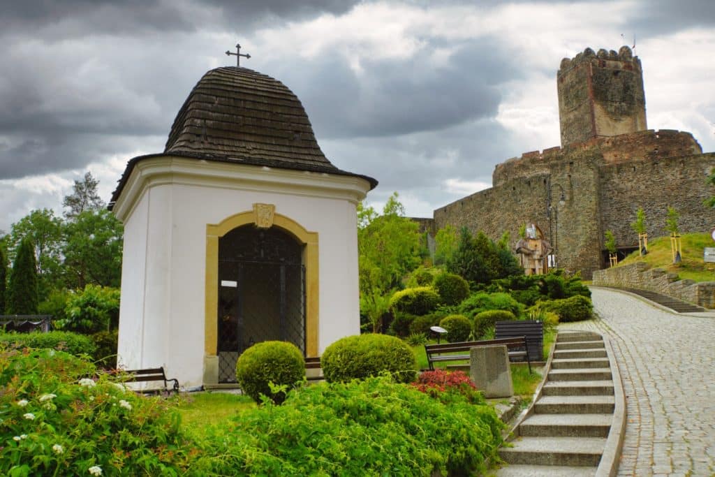Blick von unten auf eine kleine weiße Kapelle mit dunklem Schindeldach im Vordergrund und die mittelalterliche Bolkoburg in Niederschlesien auf einem Hügel im Hintergrund unter bewölktem Himmel.