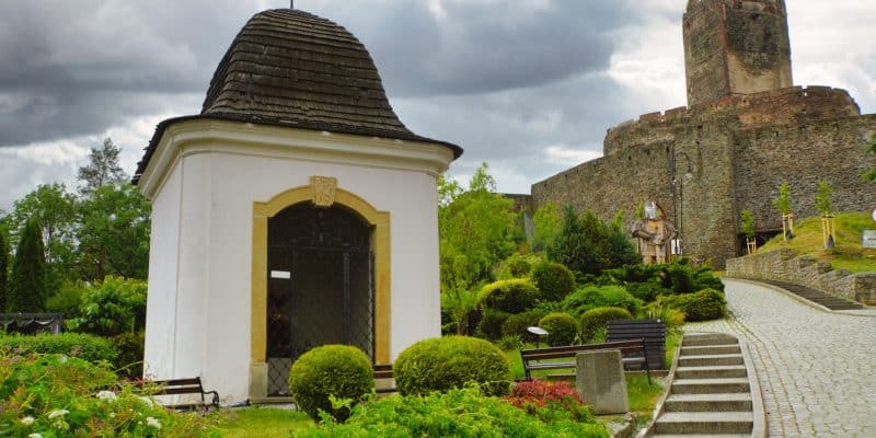 Blick von unten auf eine kleine weiße Kapelle mit dunklem Schindeldach im Vordergrund und die mittelalterliche Bolkoburg in Niederschlesien auf einem Hügel im Hintergrund unter bewölktem Himmel.