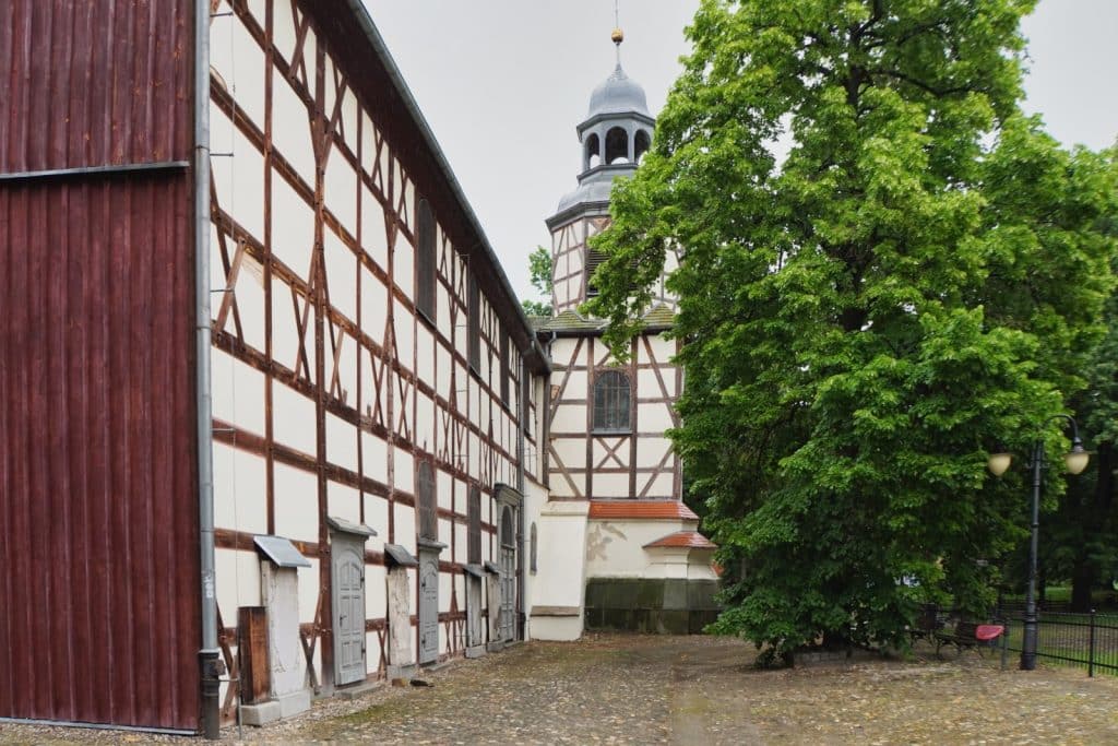 Außenansicht der Friedenskirche in Jawor, Niederschlesien, mit ihrer charakteristischen Fachwerkfassade und einem Kirchturm mit barocker Haube neben einem großen, grünen Baum.