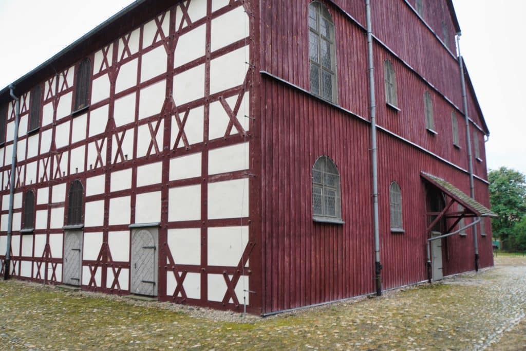 Außenansicht der Friedenskirche in Jawor, Niederschlesien, mit einer markanten Fassade aus weißem Fachwerk mit dunkelroten Holzbalken und einer angrenzenden Wand aus vertikalen roten Holzbrettern.