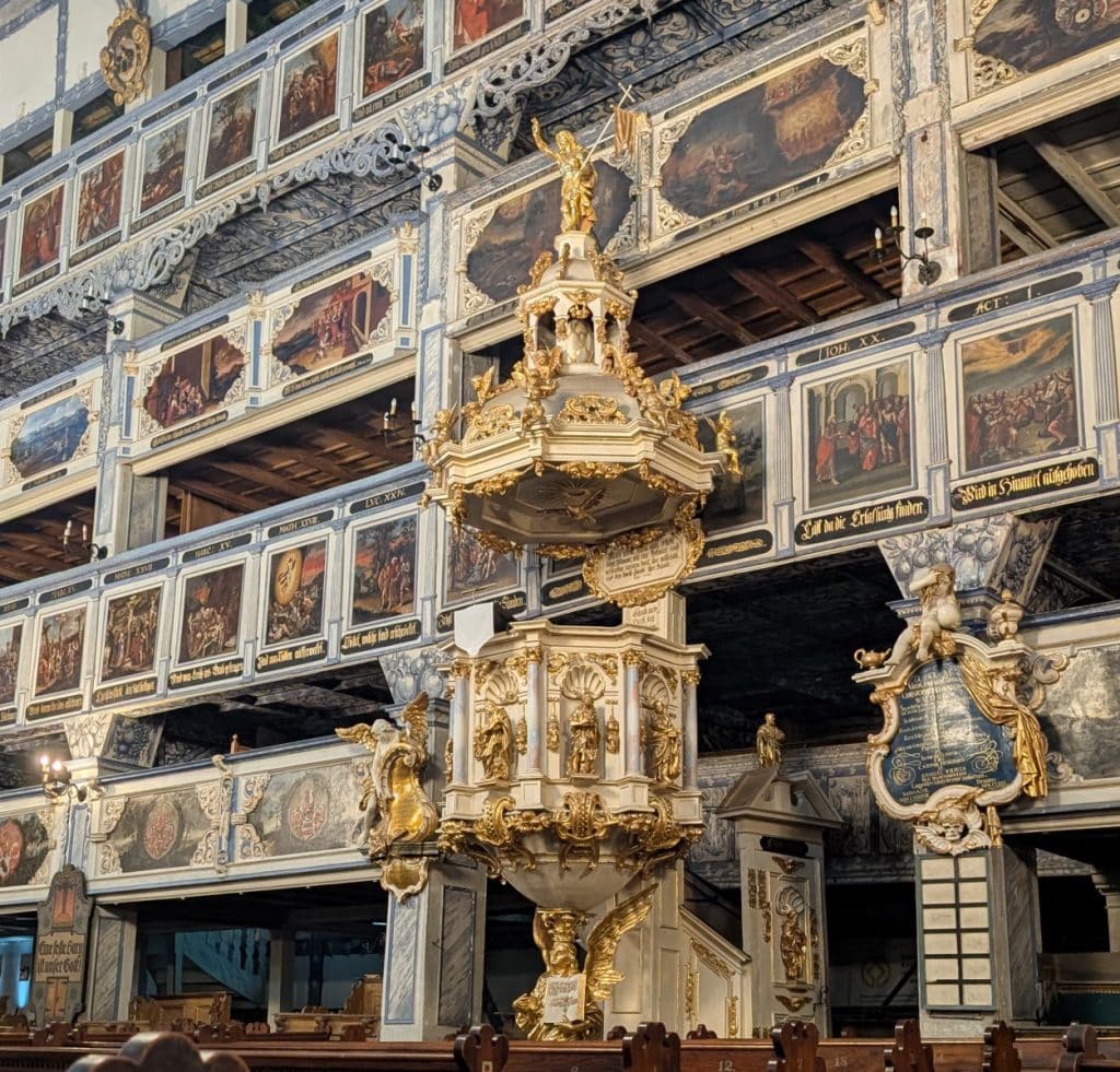 Die prunkvolle, weiß-goldene Barockkanzel der Friedenskirche in Jawor, Niederschlesien, ist umgeben von reich verzierten Emporen mit zahlreichen biblischen Gemälden und Inschriften.