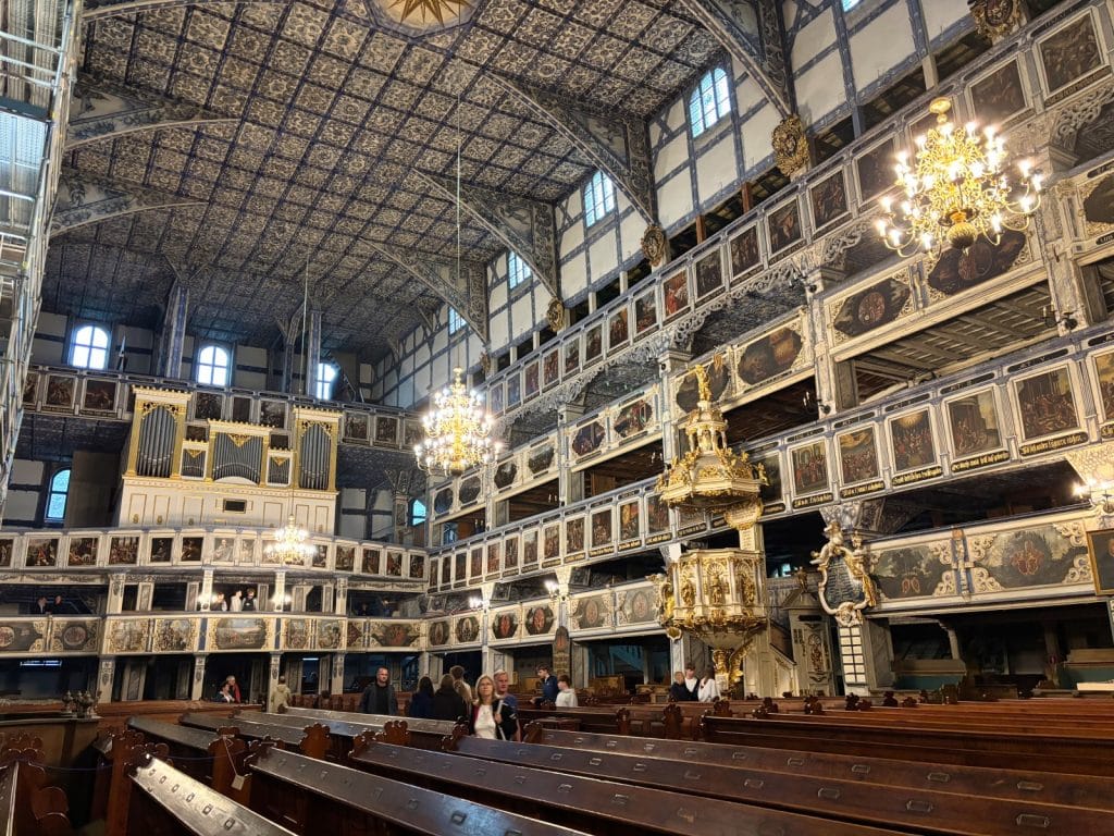 Das prachtvolle Kirchenschiff der Friedenskirche in Jawor, Niederschlesien, beeindruckt durch seine mehrstöckigen, reich mit biblischen Gemälden verzierten Holzgalerien, eine goldene Kanzel und die kunstvoll gemusterte Decke. Eine Gruppe von Besuchern steht zwischen den dunklen Holzbänken unter den hängenden Kronleuchtern und der großen Orgel.