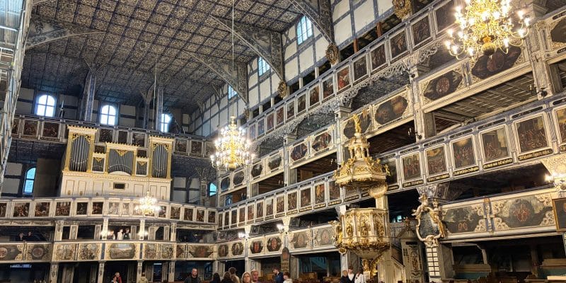 Das prachtvolle Kirchenschiff der Friedenskirche in Jawor, Niederschlesien, beeindruckt durch seine mehrstöckigen, reich mit biblischen Gemälden verzierten Holzgalerien, eine goldene Kanzel und die kunstvoll gemusterte Decke. Eine Gruppe von Besuchern steht zwischen den dunklen Holzbänken unter den hängenden Kronleuchtern und der großen Orgel.