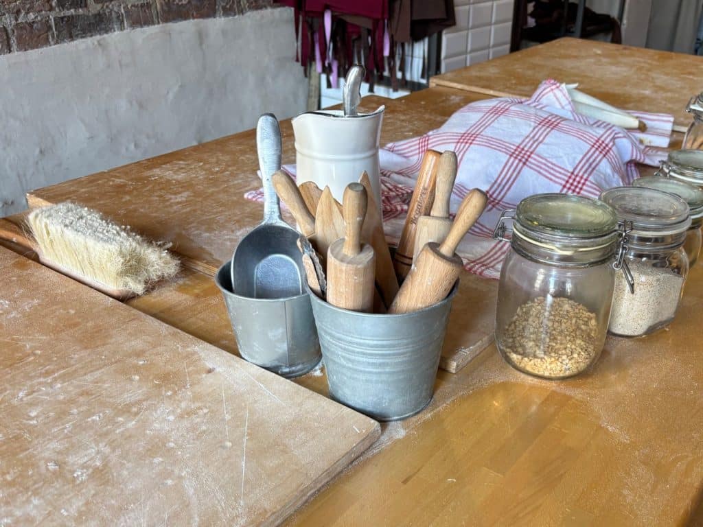 Ein bemehlter Backplatz zur Herstellung von Posener Martinshörnchen mit verschiedenen Nudelhölzern in einem Metalleimer, Teigschabern, einer Bürste und Vorratsgläsern auf einem Holztisch.