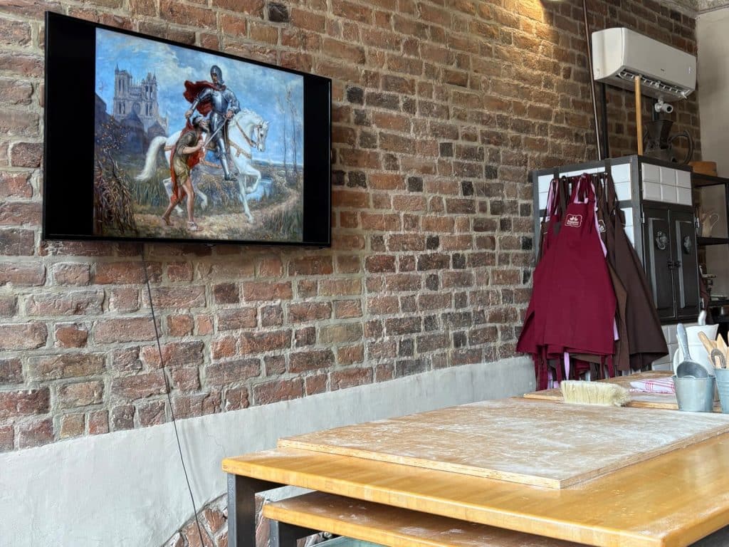 Ein Fernseher an einer Ziegelwand zeigt ein Gemälde der Geschichte des heiligen Martin, wie er seinen Mantel teilt, während im Vordergrund bemehlte Arbeitstische für die Zubereitung von Martinshörnchen bereitstehen.