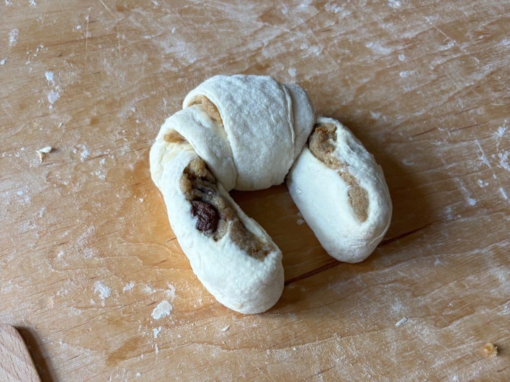 Ein ungebackenes Posener Martinshörnchen aus hellem Teig mit sichtbarer Nuss-Rosinen-Füllung liegt auf einer bemehlten Holzoberfläche.