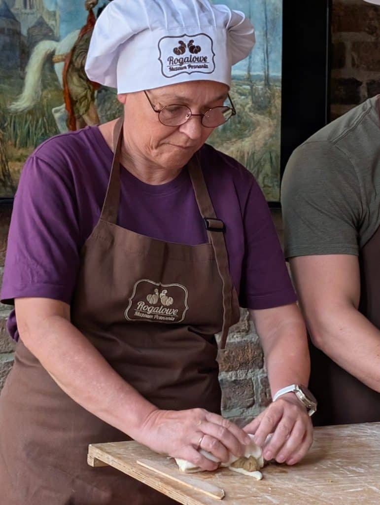 Eine Frau mit Kochmütze und Schürze formt konzentriert ein traditionelles Posener Martinshörnchen aus Teig auf einer bemehlten Holzfläche.