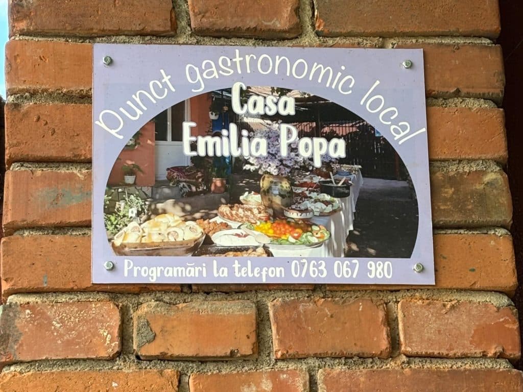 Ein Schild an einer Ziegelwand weist auf den lokalen gastronomischen Punkt „Casa Emilia Popa“ in der Region Timiș hin, illustriert mit einem Foto eines reich gedeckten Buffettisches im Freien und einer Telefonnummer für Reservierungen.
