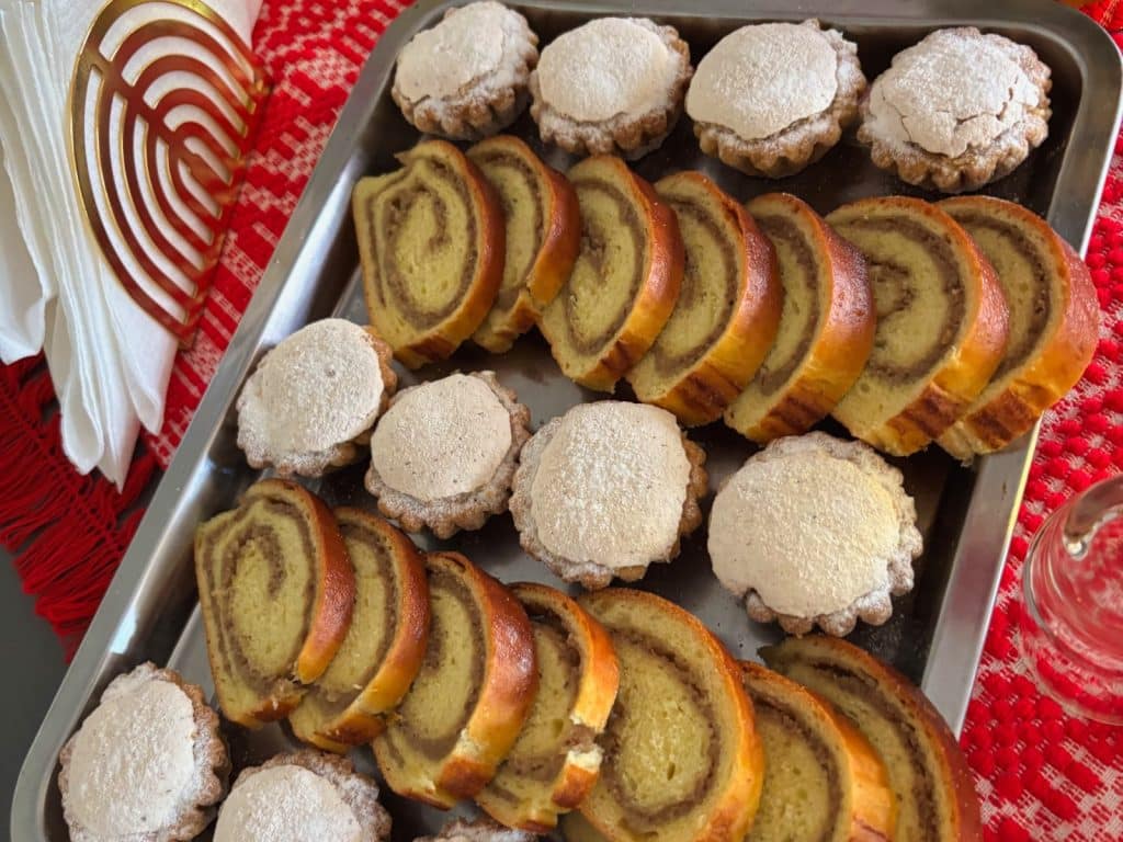 Ein silbernes Tablett ist reichlich mit traditionellem Gebäck aus der Region Timiș gefüllt, bestehend aus abwechselnden Reihen von goldbraunen Nussrollen-Scheiben und runden, mit Puderzucker bestäubten Plätzchen. Das Gebäck liegt auf einer rot-weiß gemusterten Tischdecke neben weißen Servietten in einem dekorativen Halter.