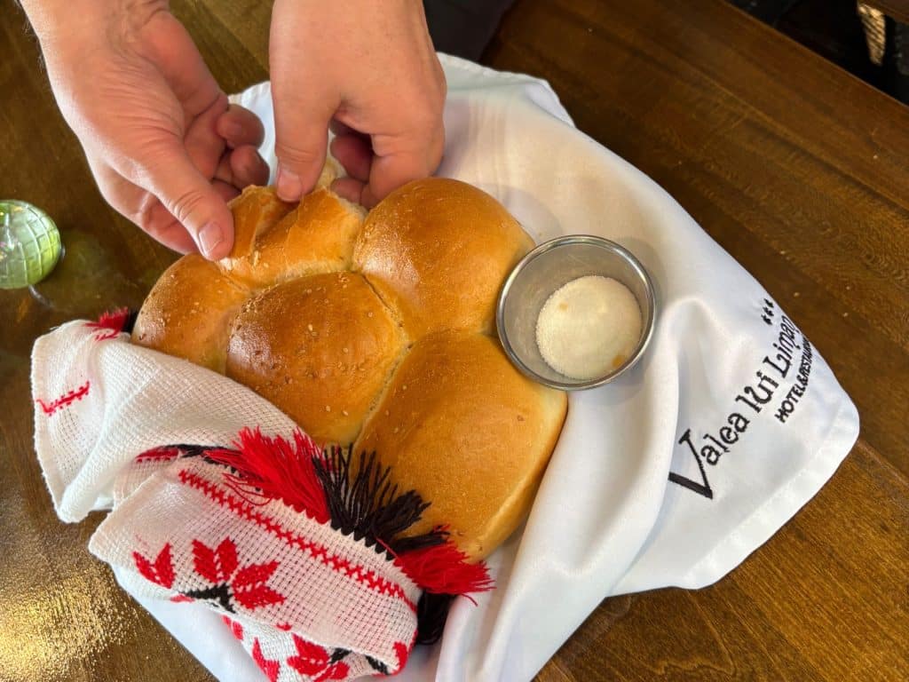 Zwei Hände brechen ein Stück von einem traditionellen Brot ab, das zusammen mit einem Schälchen Salz in einem bestickten Tuch im Restaurant Valea lui Liman in der Region Timiș serviert wird.