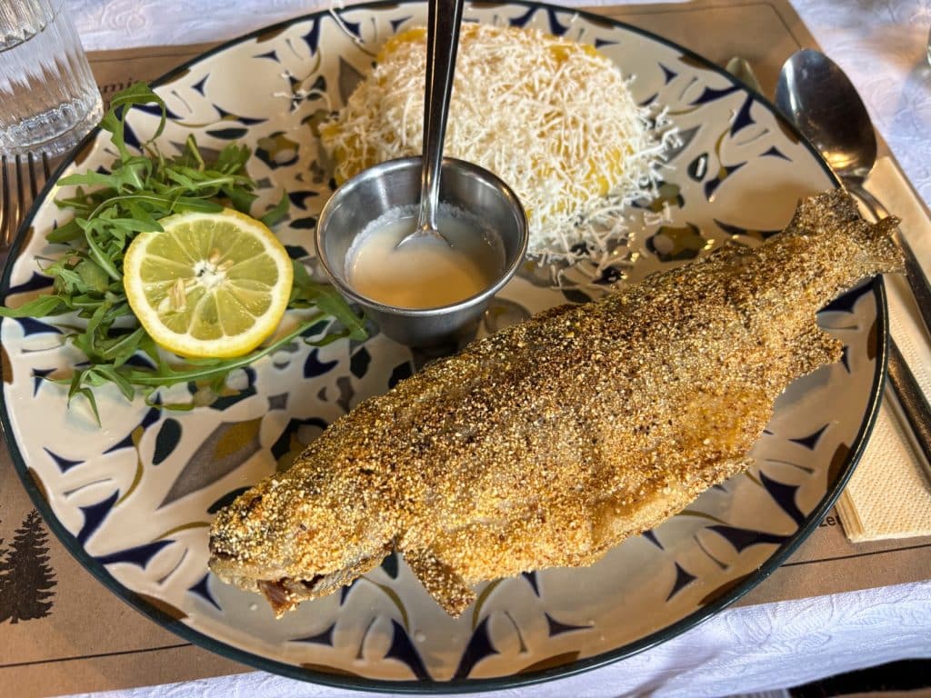 Ein in Maismehl gewendeter, gebratener ganzer Fisch wird in einem Fischrestaurant in der Region Timis zusammen mit Polenta mit Käse, einer hellen Sauce, Rucola und einer Zitronenscheibe auf einem gemusterten Teller serviert.