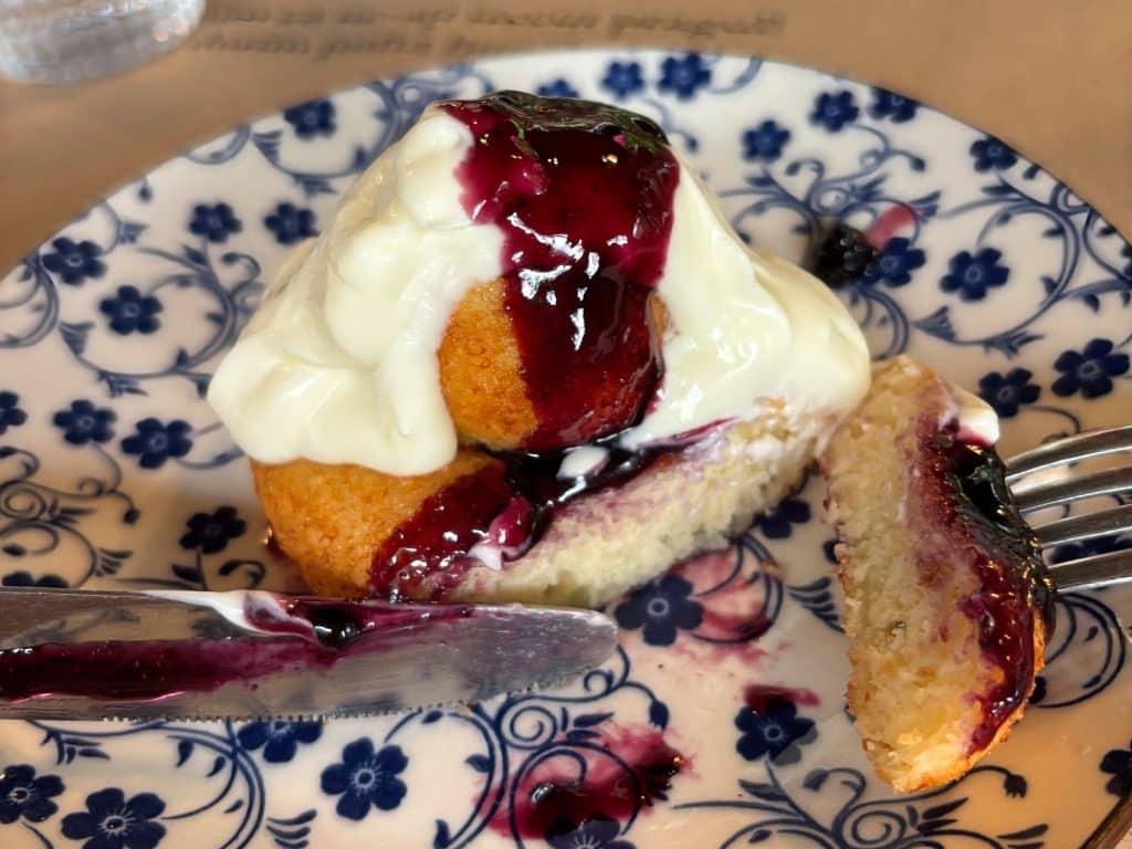 Ein angeschnittener rumänischer Papanasi-Donut, serviert in einem Fischrestaurant in der Region Timiș, ist mit weißer Sauerrahm und dunkler Blaubeermarmelade auf einem blau-weiß gemusterten Teller angerichtet.