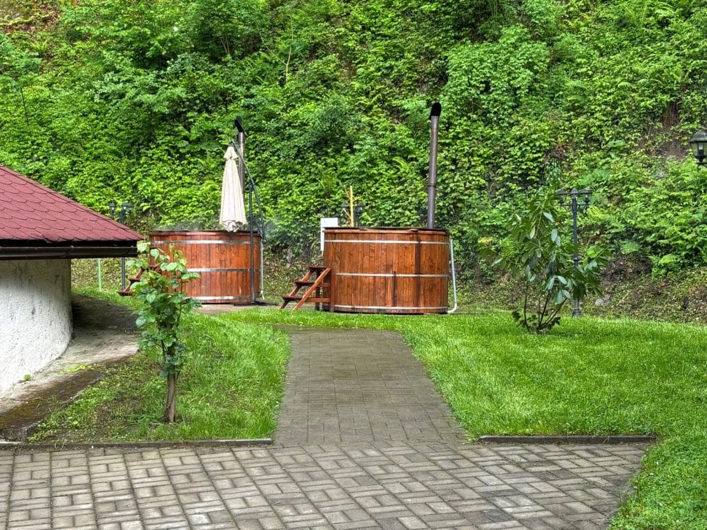 Zwei hölzerne Salzwassertonnen stehen auf einer grünen Wiese vor einer bewaldeten Böschung bei einem Fischrestaurant in der Region Timiș. Ein gepflasterter Weg führt zu den Badefässern, die mit Schornsteinen und einer kleinen Holztreppe ausgestattet sind.