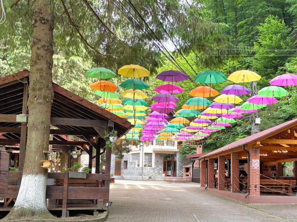 Eine gepflasterte Terrasse im Complexul Turistic Valea lui Liman in der Region Timis, über der zahlreiche bunte Regenschirme an Drähten zwischen bewaldeten Hängen und rustikalen Holzgebäuden schweben.