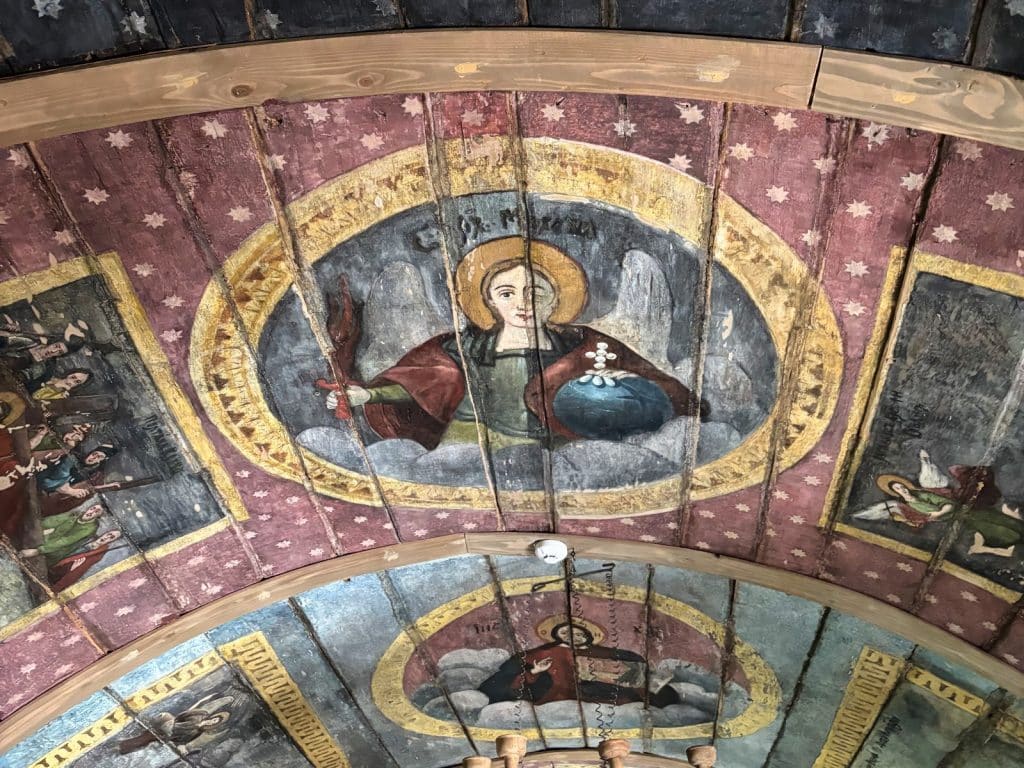 Sehenswürdigkeiten Kreis Timis : Die bemalte Holzdecke einer Kirche in der Region Timiș zeigt religiöse Ikonen auf gewölbten Holzpaneelen, darunter eine zentrale Darstellung eines Engels mit Heiligenschein, Schwert und Reichsapfel vor einem sternenbesetzten Hintergrund.