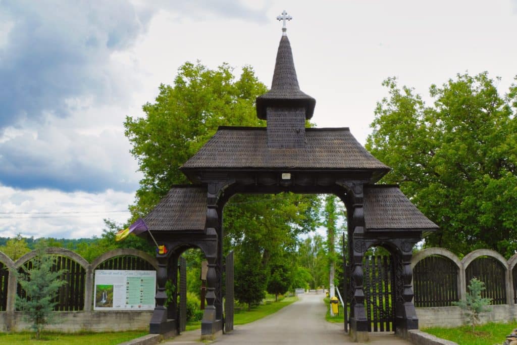 Ein großes, traditionelles Eingangstor aus dunklem Holz mit drei spitzen Schindeldächern und einem Kreuz führt zum Kloster in der Region Timiș.links neben der Einfahrt befindet sich ein Zaun mit einer Informationstafel und einer rumänischen Flagge.