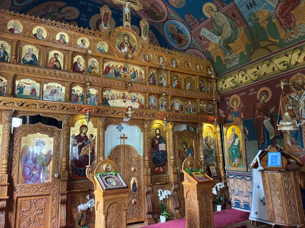 Sehenswürdigkeiten Kreis Timis : Das reich verzierte Innere einer Kapelle im Kloster der Region Timis zeigt eine goldene Ikonostase mit zahlreichen Heiligenbildern und kunstvollen Schnitzereien vor farbenfroh bemalten Wänden und Decken. Vor der Trennwand stehen hölzerne Pulte mit Ikonen und weißen Blumenarrangements auf einem roten Teppich.