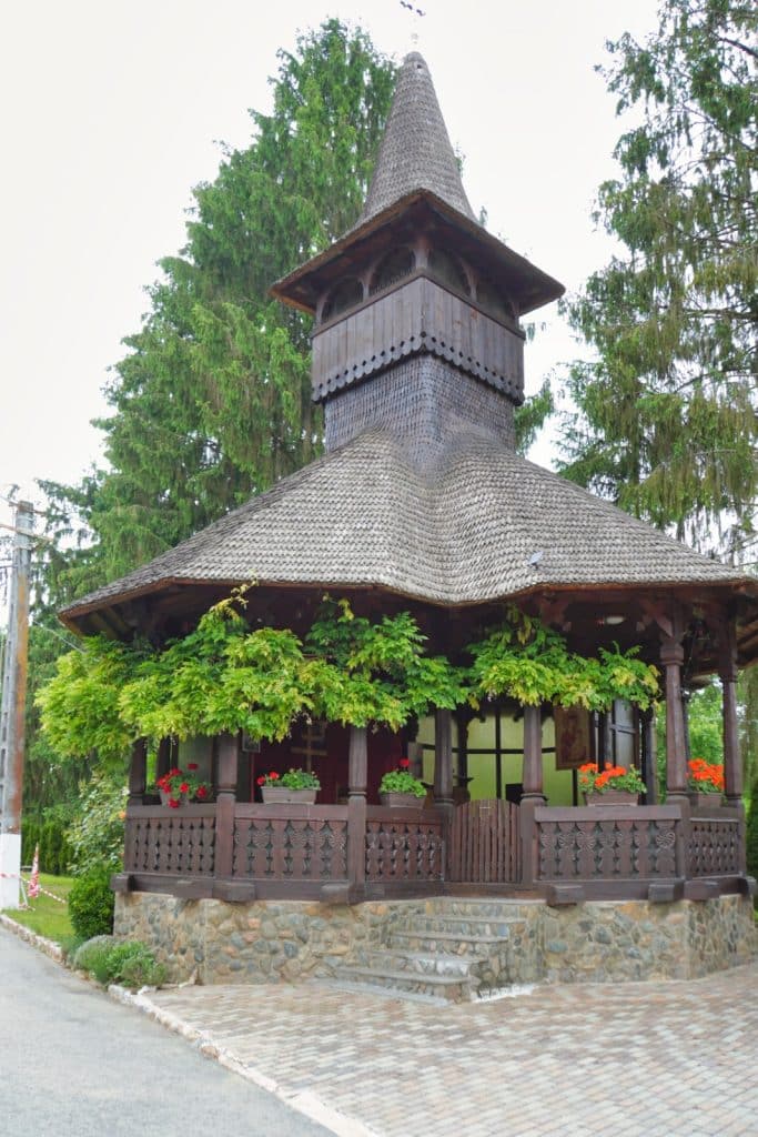 Eine hölzerne Sommerkirche im Kloster der Region Timiș mit einem markanten Schindeldach, einem spitzen Turm und einer mit Blumenkästen sowie grünen Ranken geschmückten Veranda auf einem Steinsockel.