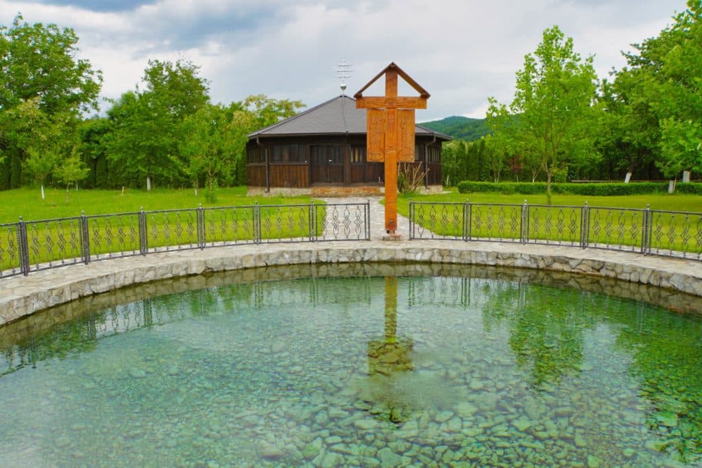 Ein hölzernes Kreuz steht am Rand einer kreisförmig eingefassten warmen Quelle im Kloster der Region Timis, in deren klarem Wasser sich das Kreuz und ein dunkles Holzgebäude im Hintergrund spiegeln.