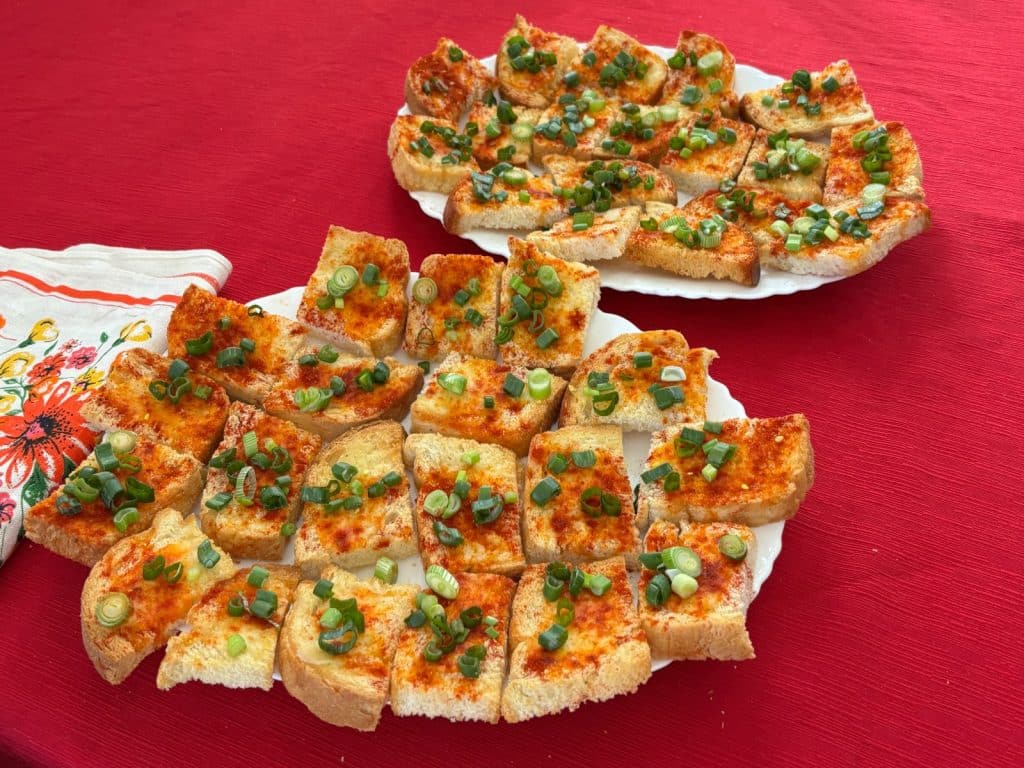 Zwei weiße Teller mit gerösteten Scheiben von Region Timis Mühle Brot, die mit Paprikapulver gewürzt und mit frisch geschnittenen Frühlingszwiebeln bestreut sind, stehen auf einer roten Tischdecke.