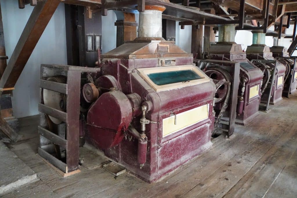 Innenansicht einer historischen Mühle in der Region Timiș mit einer Reihe von großen, weinroten Mahlmaschinen aus Holz und Metall auf einem rustikalen Dielenboden.