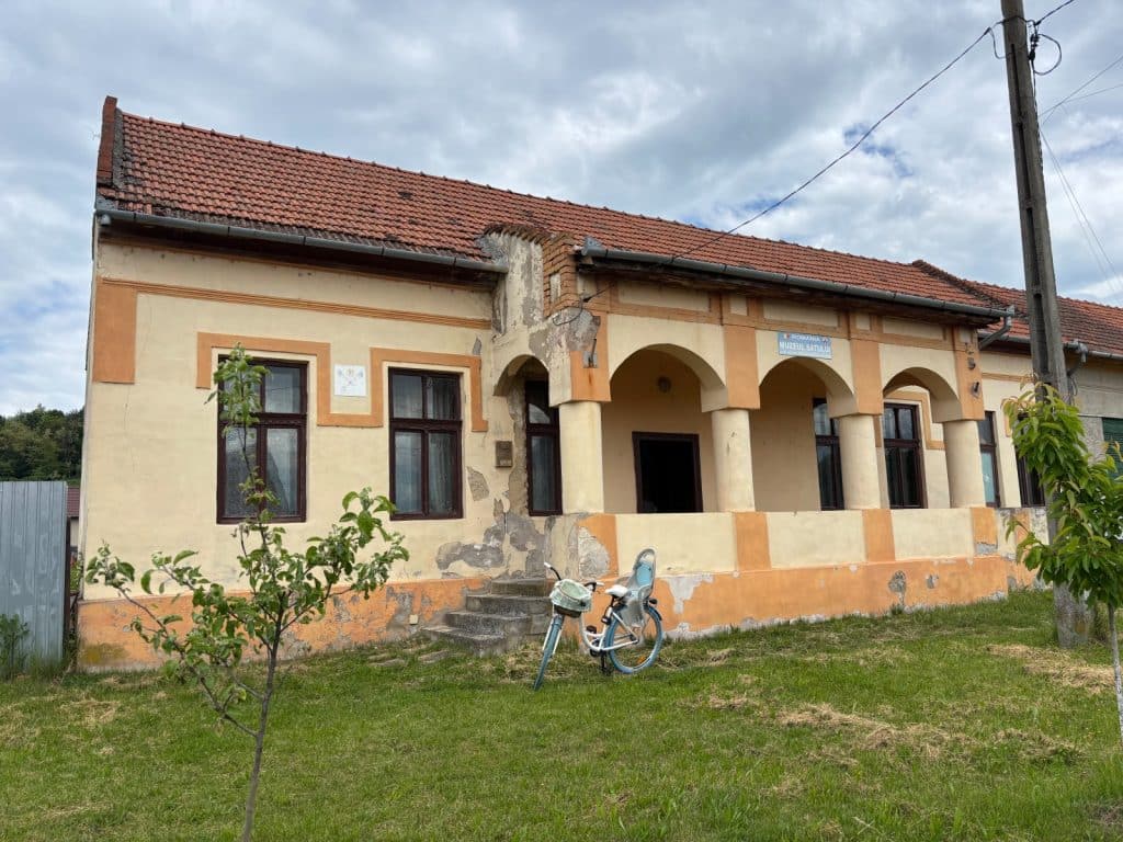 Sehenswürdigkeiten Kreis Timis : Ein einstöckiges, hellgelbes Gebäude des Dorfmuseums in der Region Timiș mit einer markanten Säulenvorhalle, einem roten Ziegeldach und einem davor parkenden hellblauen Fahrrad auf einer Wiese.