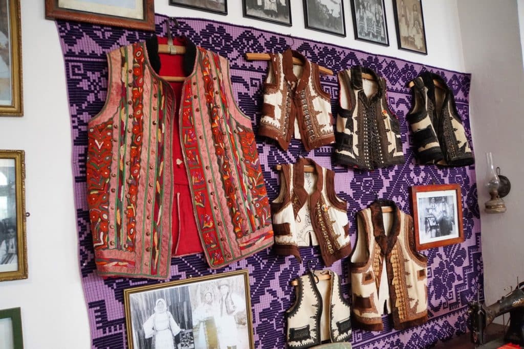 Eine Sammlung traditioneller rumänischer Trachtenwesten aus der Region Timiș hängt zusammen mit historischen Schwarz-Weiß-Fotografien an einer Wand vor einem violett gemusterten Wandteppich in einem Museum.