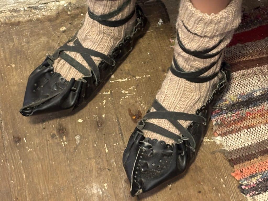 Ein Paar traditionelle schwarze Lederschuhe aus dem Region Timiș Museum, die mit dunklen Riemen über dicken, hellen Strickstrümpfen geschnürt sind.