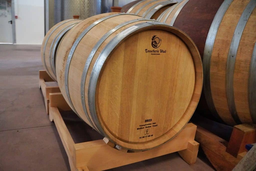 In einem Weingut in der Region Timiș lagern mehrere Weinfässer aus hellem Eichenholz auf hölzernen Gestellen, wobei das vordere Fass die Aufschrift „Tonnellerie Vlad“ und das Jahr 2023 trägt.