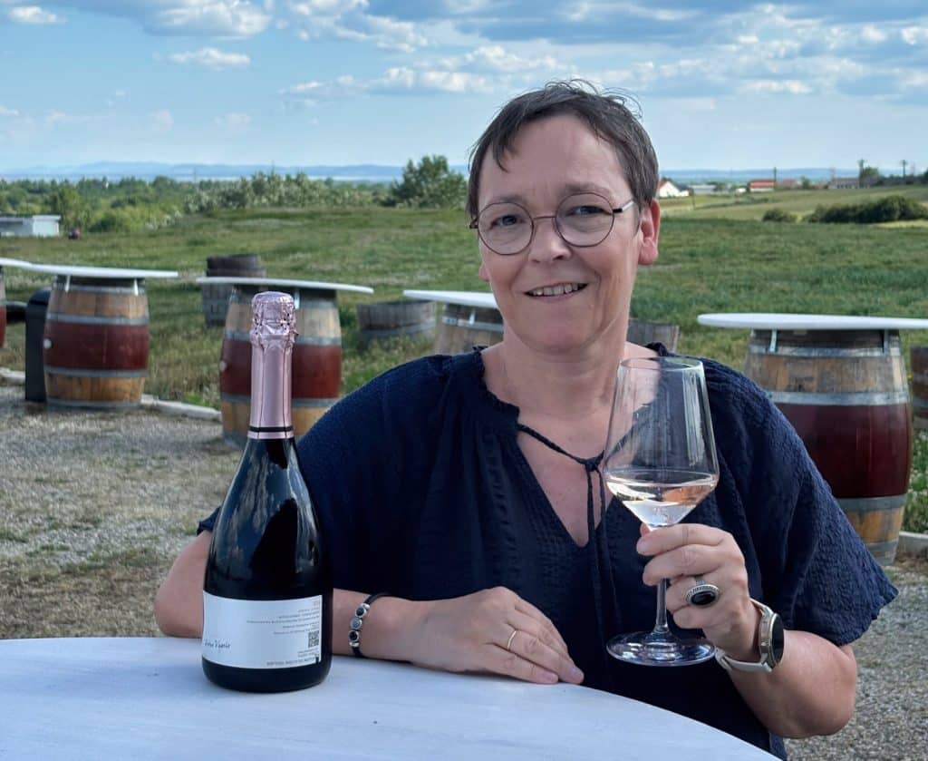 Eine lächelnde Frau mit Brille sitzt an einem Tisch auf einem Weingut in der Region Timiș und prostet mit einem Glas Roséwein neben einer passenden Flasche Sekt zu. Im Hintergrund erstreckt sich eine weite Landschaft mit Weinfässern, die als Stehtische dienen, unter einem leicht bewölkten Himmel.