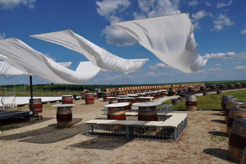 Eine sonnige Terrasse eines Weinguts in der Region Timis mit zahlreichen Weinfässern, die als Stehtische dienen, und weißen, im Wind wehenden Sonnensegeln unter blauem Himmel.