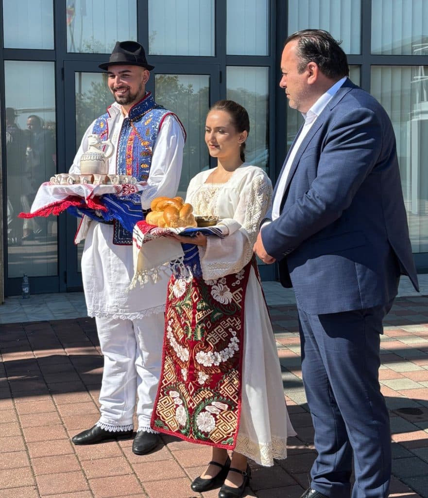 Ein Mann und eine Frau in traditionellen rumänischen Trachten begrüßen einen Mann im Anzug mit Brot, Salz und einem Krug auf einem Tablett.