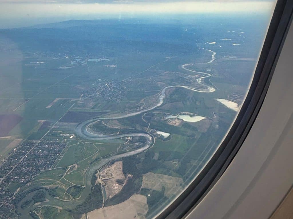 Blick aus einem Flugzeugfenster auf die rumänische Landschaft mit einem stark mäandrierenden Fluss, der sich durch grüne Felder, kleine Ortschaften und Wälder zieht. Im Hintergrund erstrecken sich unter einem leicht bewölkten Himmel weite Ebenen und ferne Hügelketten.