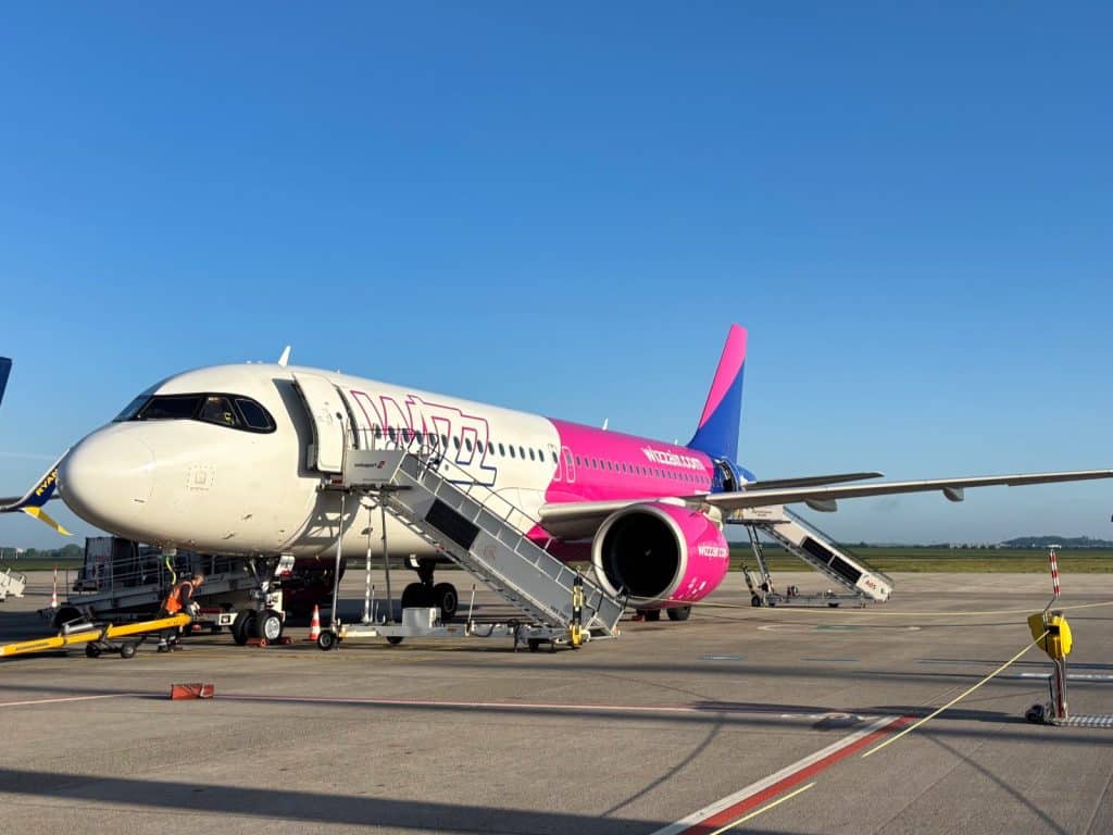 Ein weiß-pinkes Flugzeug der Fluggesellschaft Wizz Air steht bei sonnigem Wetter auf dem Rollfeld eines Flughafens in Rumänien, während Passagiertreppen an die Türen angedockt sind.