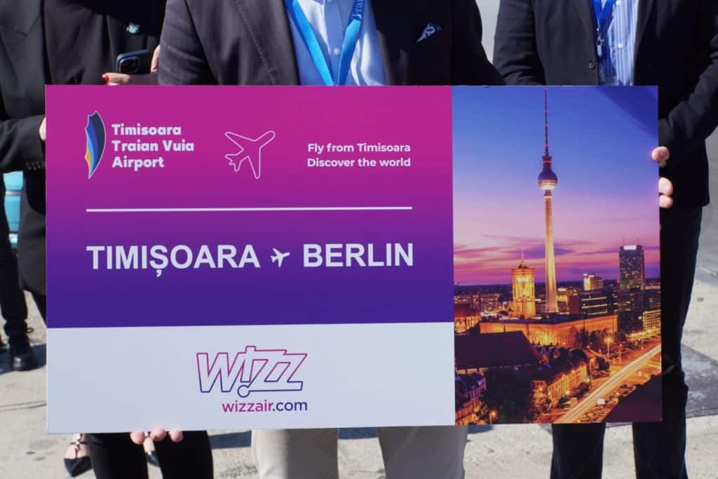 Drei Personen halten ein großes Schild, das eine neue Flugverbindung der Fluggesellschaft Wizz Air von Timișoara in Rumänien nach Berlin ankündigt, illustriert mit einem Foto des Berliner Fernsehturms bei Dämmerung.