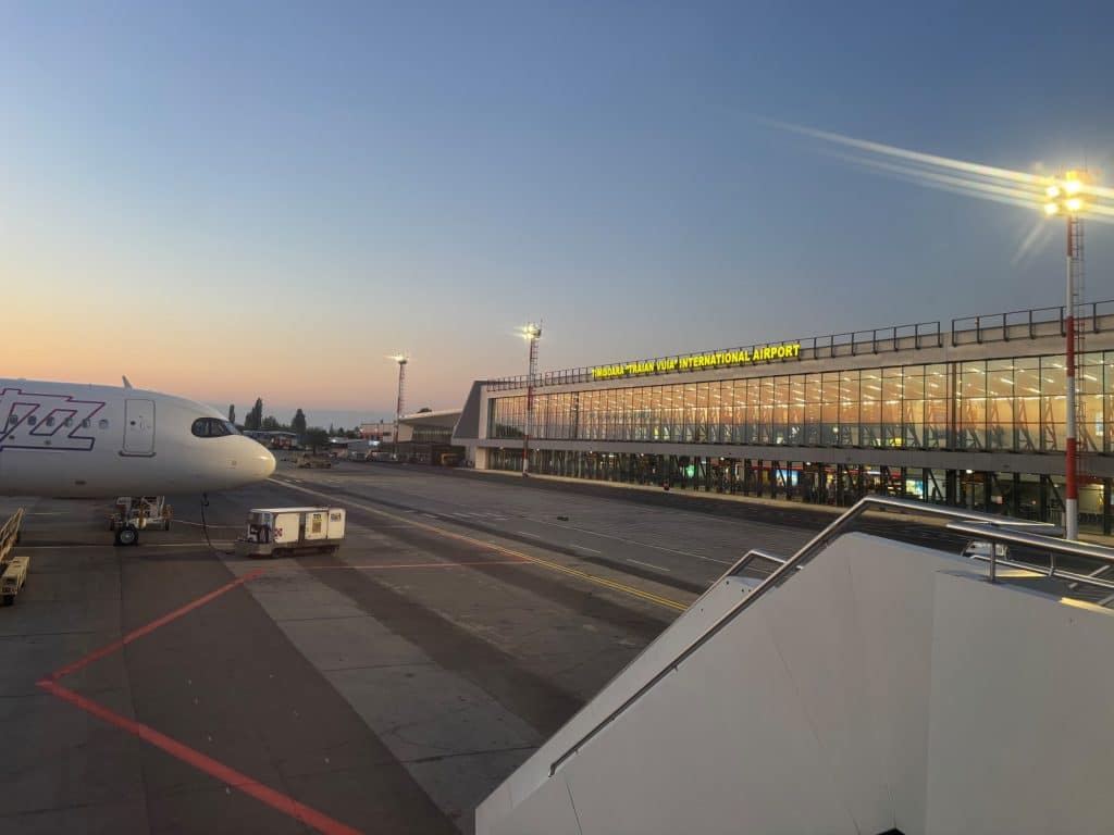 Blick vom Rollfeld auf das moderne Terminalgebäude des internationalen Flughafens „Traian Vuia“ in Timișoara, Rumänien, während der Abenddämmerung mit einem geparkten Wizz-Air-Flugzeug im Vordergrund.