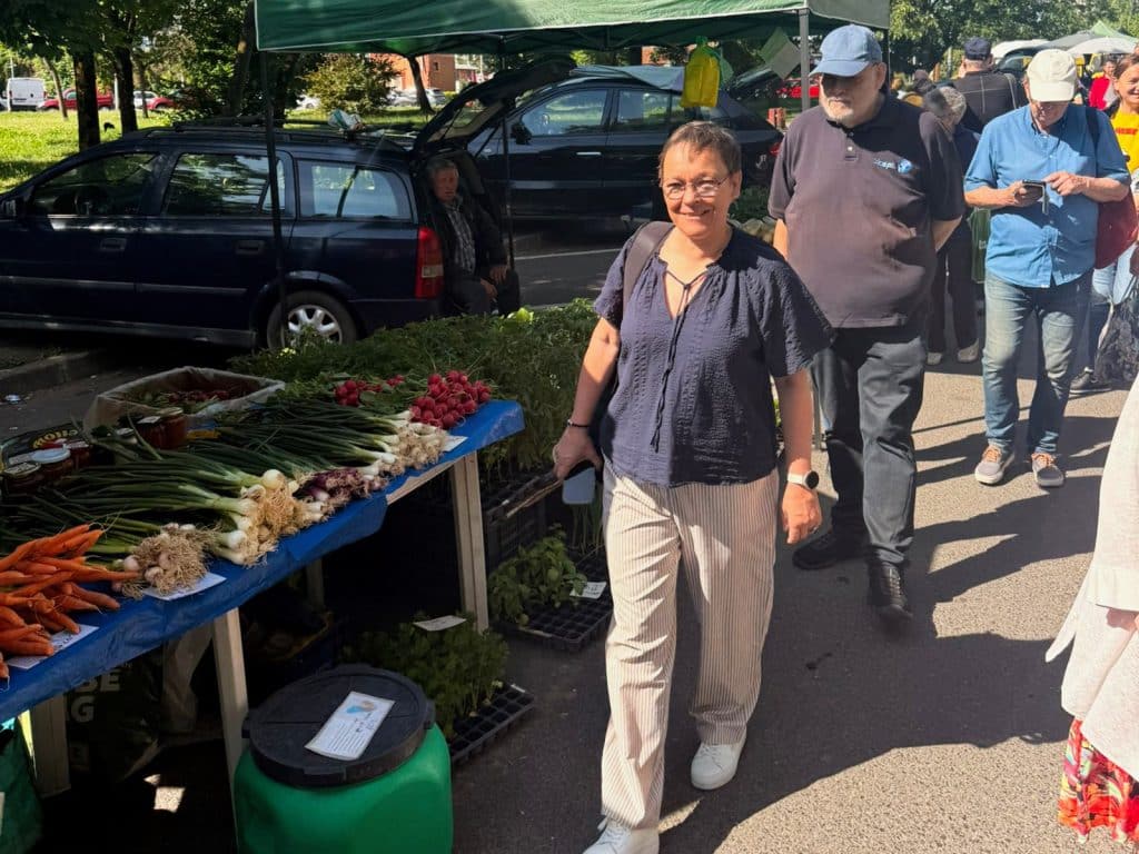Eine Frau lächelt beim Bummeln über den Timisoara Markt, während sie an einem Stand mit frischen Karotten, Frühlingszwiebeln und Radieschen vorbeiläuft. Im Hintergrund sind weitere Marktbesucher, Autos und ein grünes Marktzelt unter sonnigem Himmel zu sehen.
