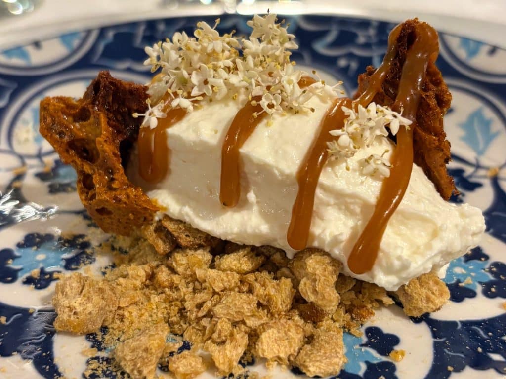 Eine kunstvoll angerichtete Nachspeise im Restaurant Casa del Sole in Timișoara, bestehend aus einer weißen Creme auf Keksbröseln, garniert mit Karamellsauce, kleinen weißen Blüten und knusprigem Waben-Toffee.