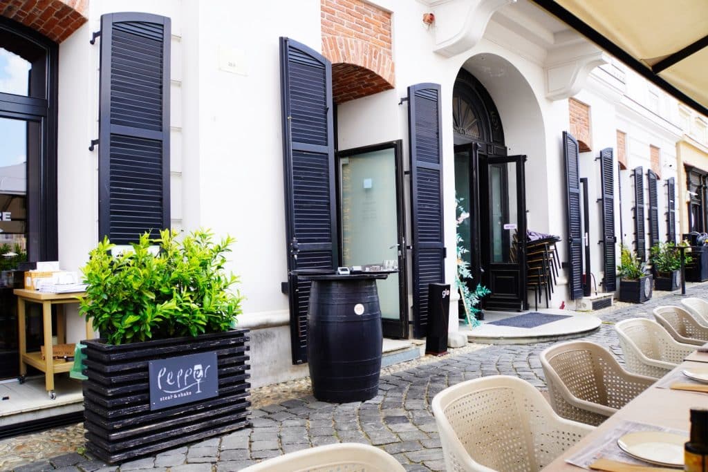 Der Eingang des Restaurants Pepper in Timișoara mit einer gepflasterten Außenterrasse, schwarzen Fensterläden an der weißen Fassade und einem Pflanzkübel im Vordergrund.
