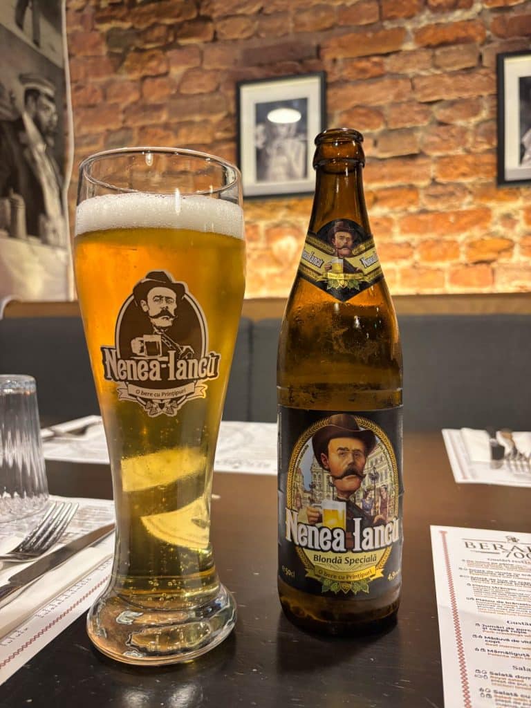 Ein frisch gezapftes Glas und eine Flasche Nenea Iancu „Blondă Specială“ Bier stehen auf einem gedeckten Tisch in einem Restaurant in Timișoara vor einer rustikalen Ziegelwand.