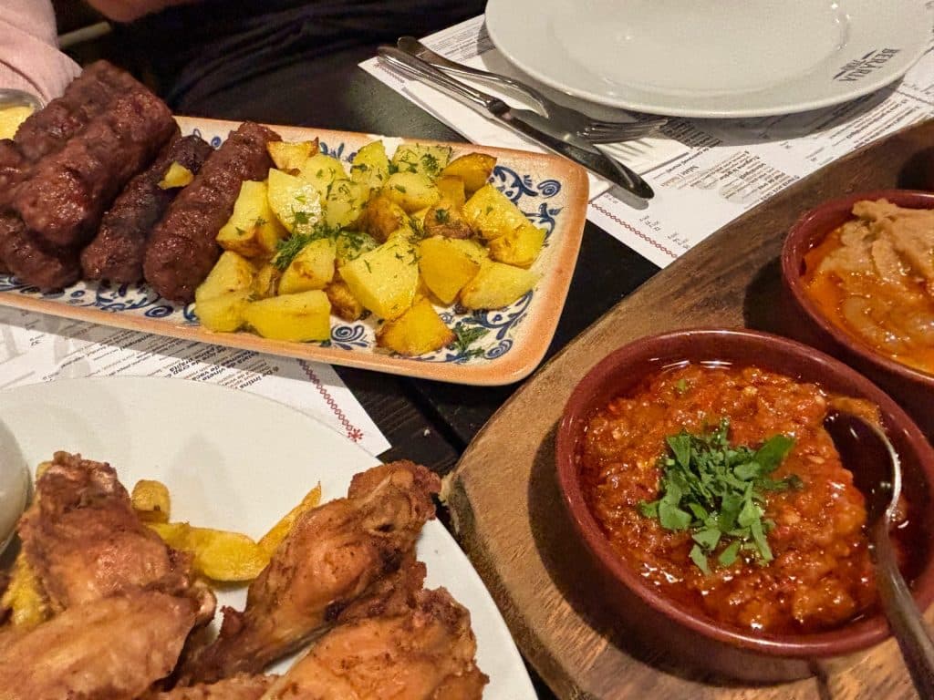 Ein reichhaltiges rumänisches Essen in einer Brauerei in Timișoara mit gegrillten Mici, Bratkartoffeln, Hähnchenflügeln und verschiedenen Beilagen wie Zacusca in Tonschalen auf einem rustikalen Holztisch.
