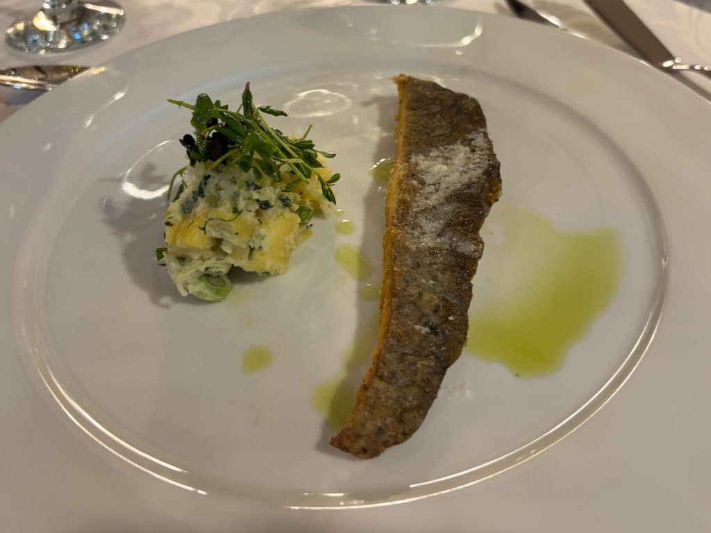 Ein gebratenes Fischfilet mit knuspriger Haut, serviert mit einem Kartoffelsalat und frischen Kräutern auf einem weißen Teller im Restaurant Casa del Sole in Timișoara.