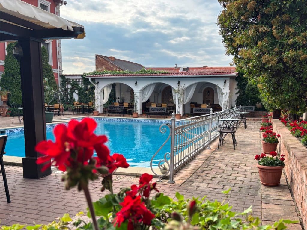Der Außenbereich des Restaurants Casa del Sole in Timișoara zeigt einen blauen Pool, der von einer gepflasterten Terrasse mit roten Blumenkästen, schmiedeeisernen Zäunen und einem weißen Gebäude mit Rundbögen umgeben ist.
