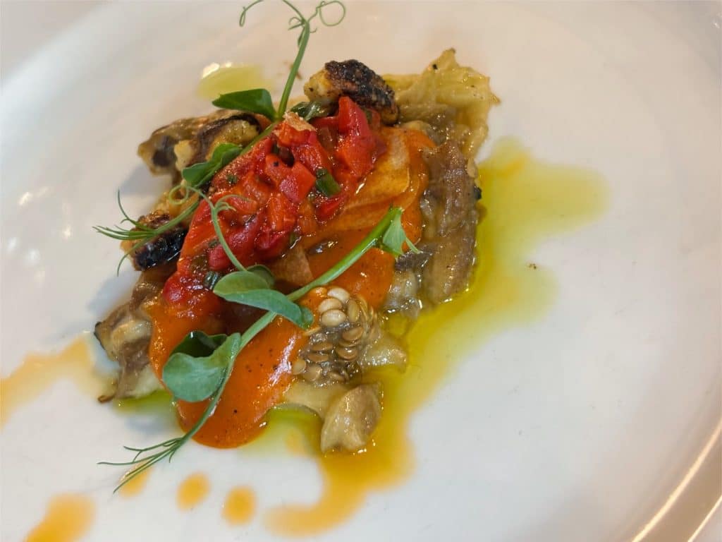 Eine kunstvoll angerichtete Vorspeise im Restaurant Casa del Sole in Timișoara, bestehend aus gegrilltem Gemüse, einer leuchtend roten Paprikasauce und frischen Kräutern auf einem weißen Teller, verfeinert mit Olivenöl.