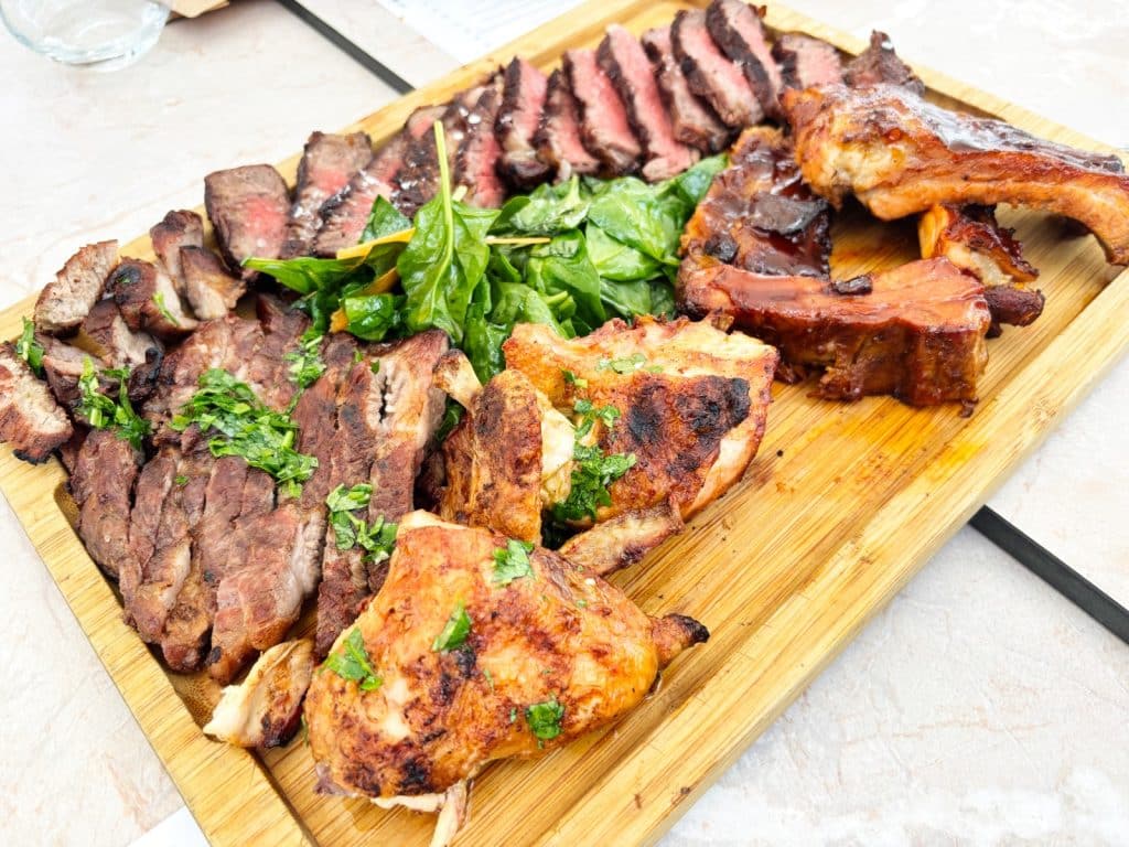 Eine reichhaltige Fleischplatte des Restaurants Pepper in Timișoara mit aufgeschnittenem Steak, gegrilltem Hähnchen, Rippchen und einem frischen Spinatsalat auf einem Holzbrett.