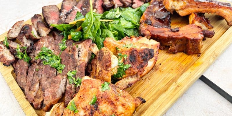 Eine reichhaltige Fleischplatte des Restaurants Pepper in Timișoara mit aufgeschnittenem Steak, gegrilltem Hähnchen, Rippchen und einem frischen Spinatsalat auf einem Holzbrett.