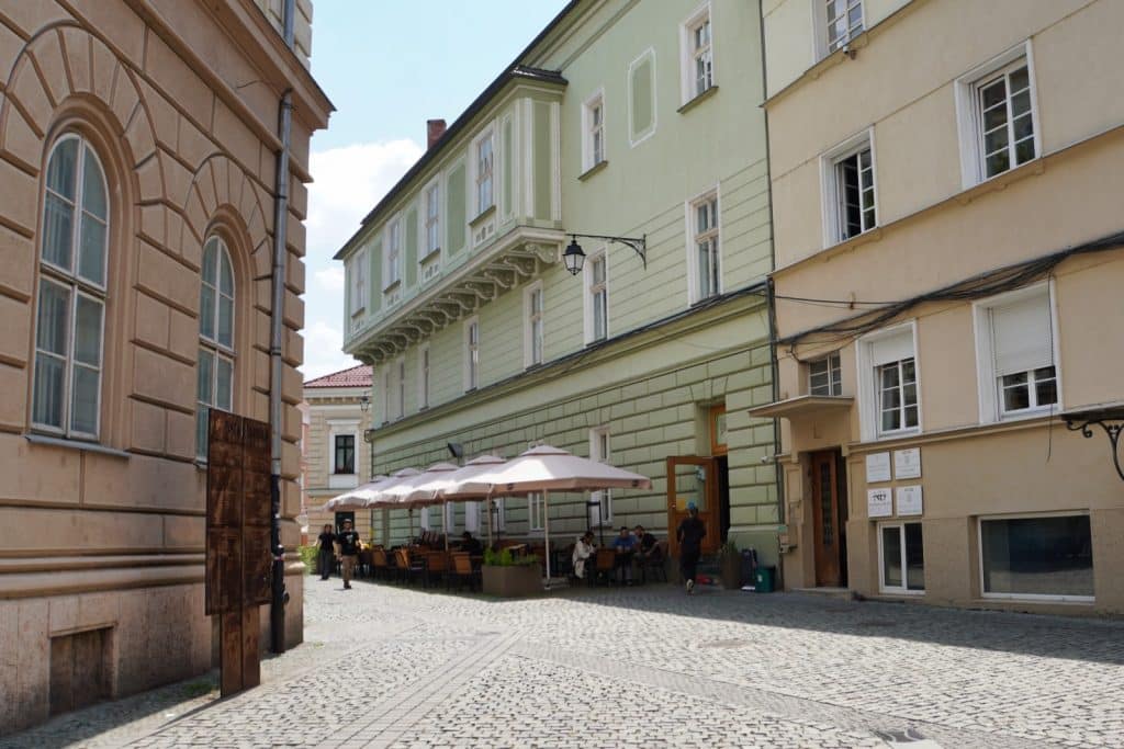 Eine gepflasterte Gasse in Timișoara mit historischen, mehrstöckigen Häusern und einem Straßencafé unter großen Sonnenschirmen, in dem Menschen an Tischen sitzen.