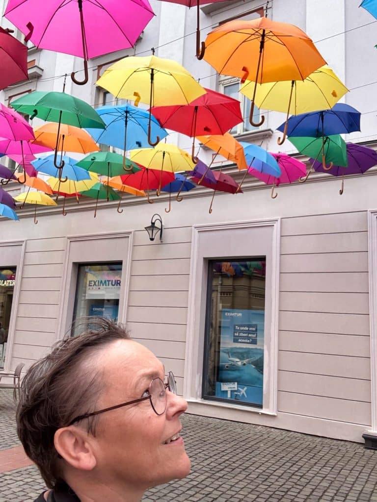 Eine Frau blickt lächelnd nach oben auf eine Vielzahl bunter Regenschirme, die in einer Straße in Timișoara über dem Kopfsteinpflaster in der Luft schweben. Im Hintergrund ist eine helle Hausfassade mit Schaufenstern zu sehen.