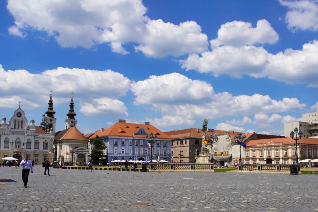 Sehenswürdigkeiten in Timișoara: Der weitläufige, gepflasterte Vereinigungsplatz in Timișoara ist von farbenfrohen historischen Gebäuden, barocken Kirchen mit markanten Türmen und Denkmälern unter einem strahlend blauen Wolkenhimmel gesäumt.