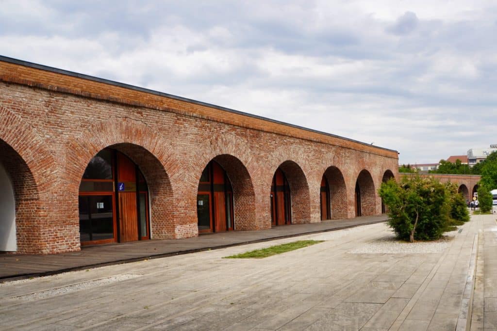 Eine Außenansicht der historischen Maria-Theresia-Bastion in Timișoara mit ihren charakteristischen Backsteinbögen, hinter denen sich moderne Glasfronten mit Holzelementen befinden. Vor dem Gebäude erstreckt sich ein gepflasterter Platz mit vereinzelten Grünflächen und kleinen Büschen unter einem bewölkten Himmel.